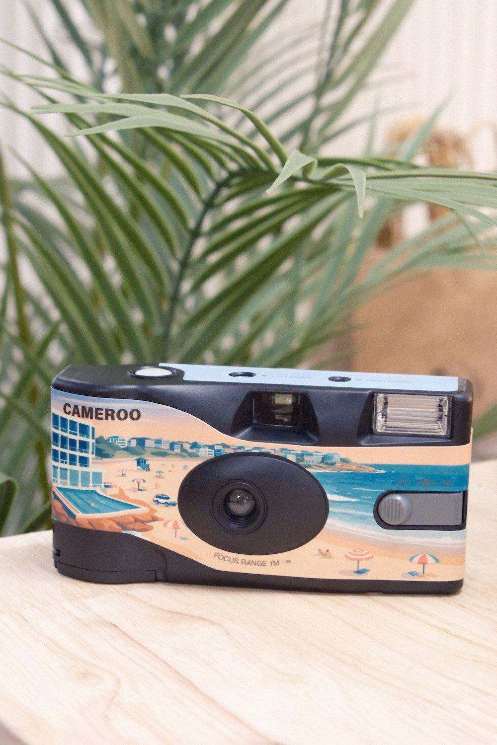 Disposable film camera with a Sydney beach illustration, blue and peach artwork, and a black lens; One Camera, 27 Frames