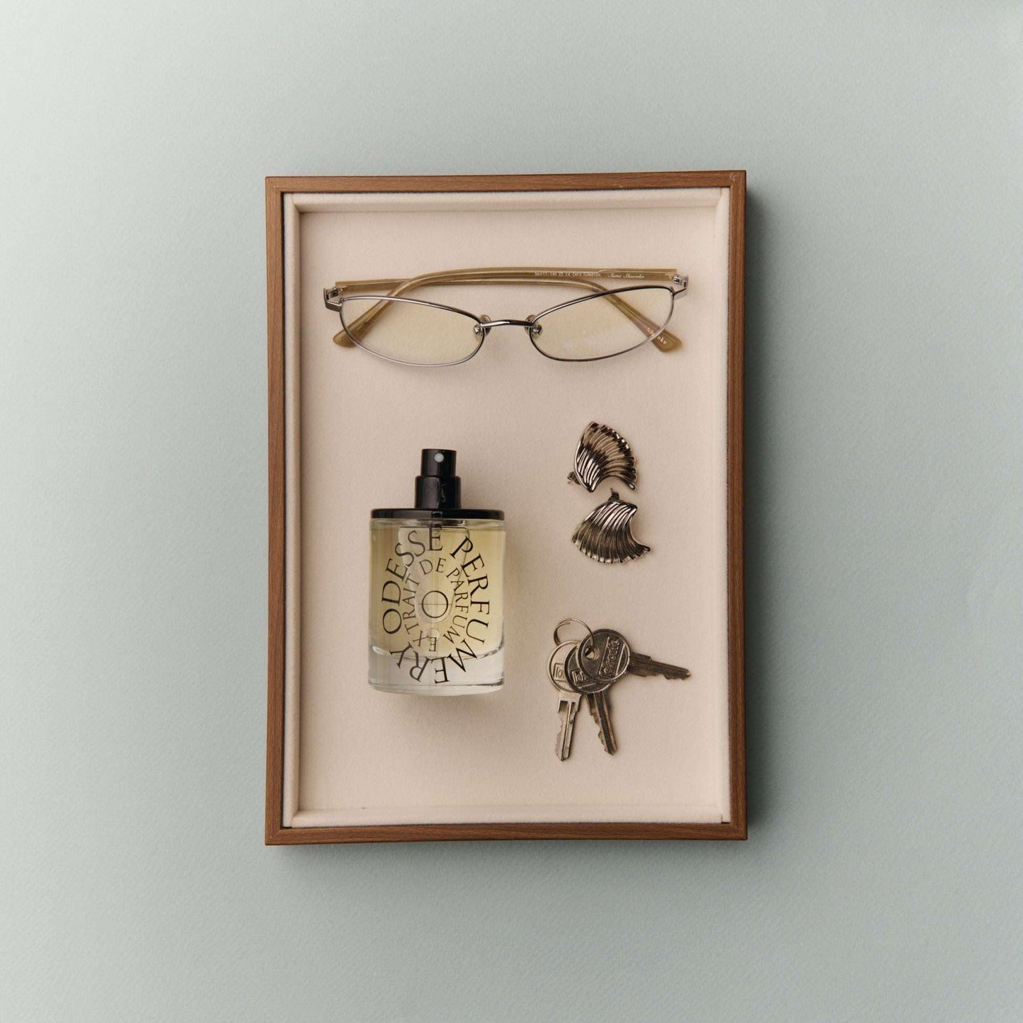 Odesse Extrait De Parfum displayed in velvet shadow box with gold-rimmed glasses, seashell earrings, and vintage keys