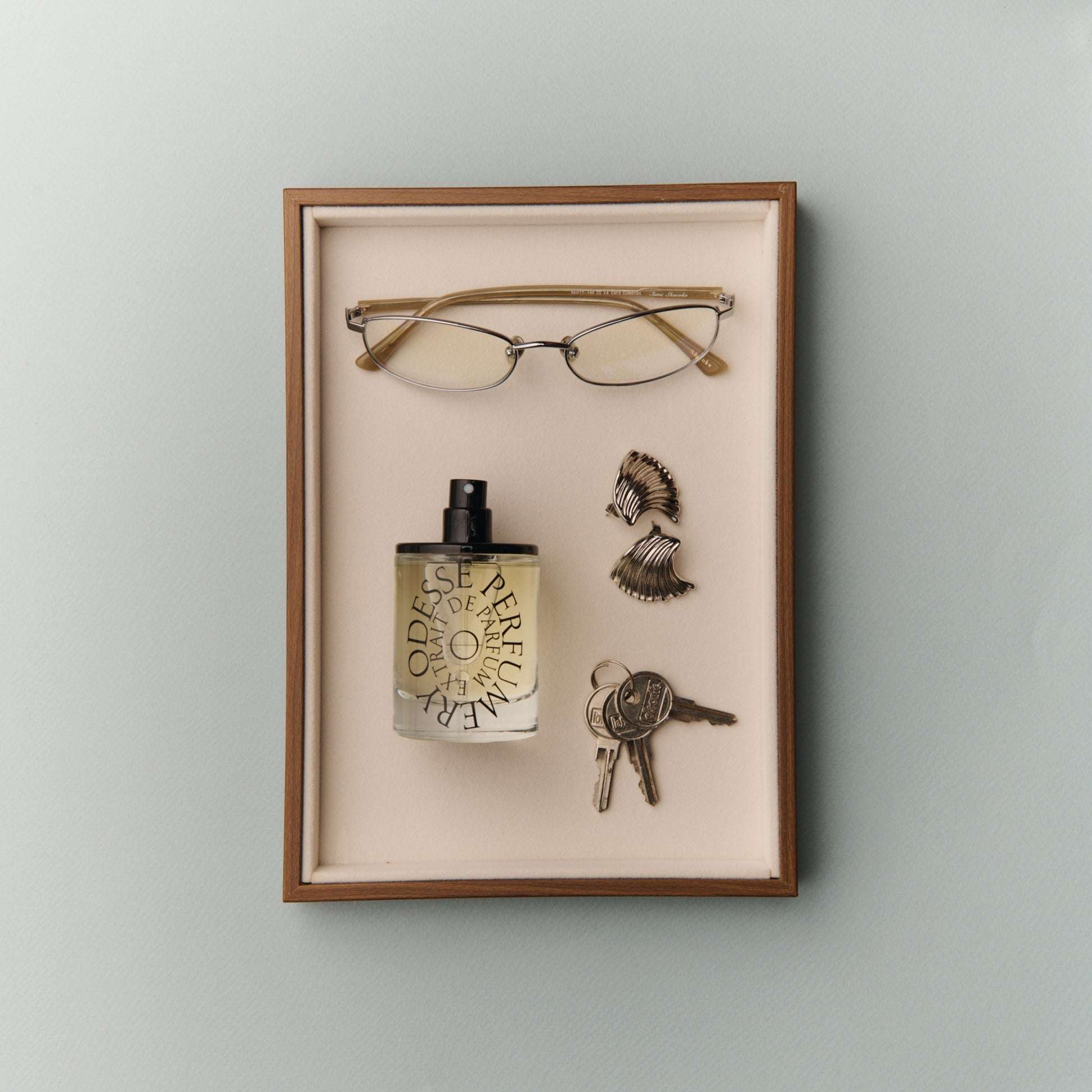 Odesse Extrait De Parfum displayed in velvet shadow box with gold-rimmed glasses, seashell earrings, and vintage keys