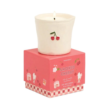 Lit Paddywax Smoked Cherry soy wax candle in embossed ceramic vessel atop illustrated retail packaging