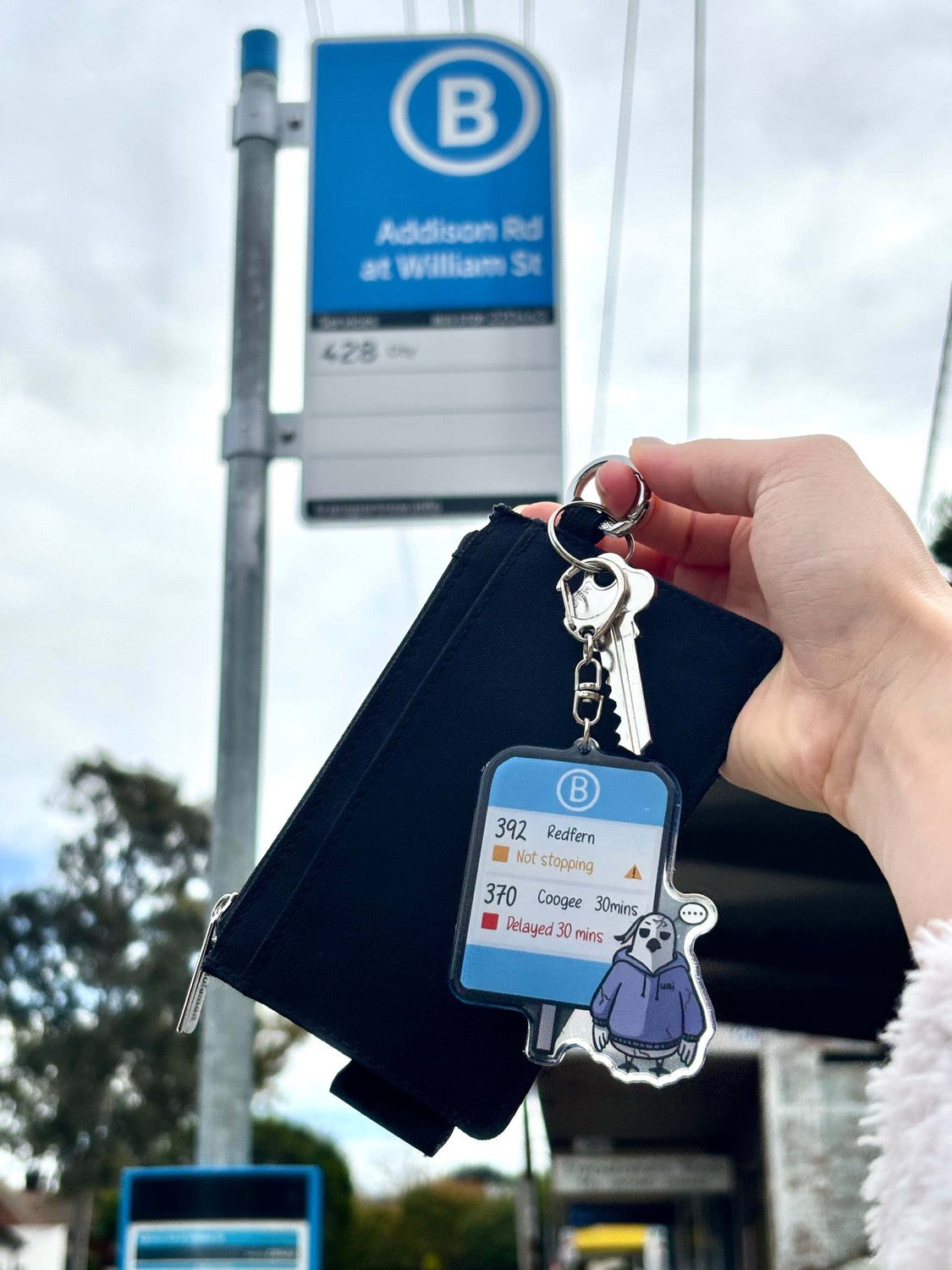 9to5 Studios keychain attached to keys at outdoor bus stop showing commuter humor design