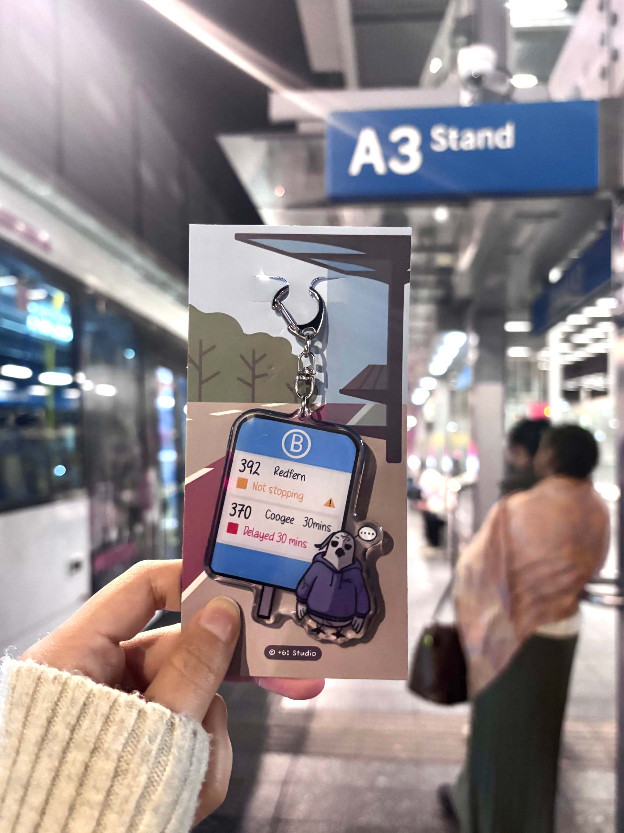 +61 Studio transit-themed keychain held against blurred station background