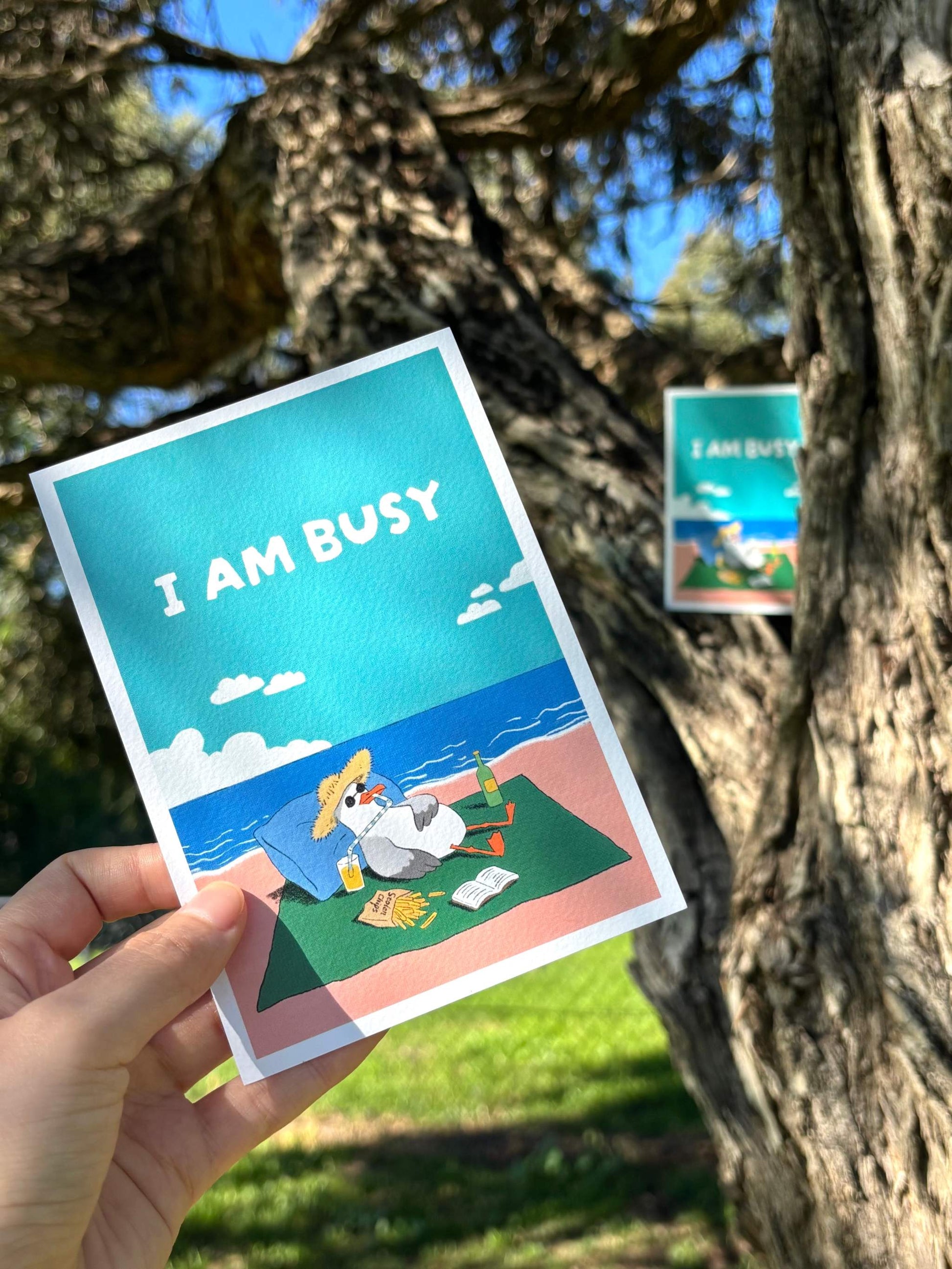 9to5 Studios +61 Studio I AM BUSY postcard with cartoon seagull in straw hat holding yellow drink on green towel