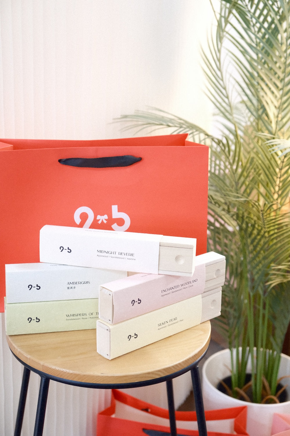 Assorted incense boxes fanned on stool beside coral-red gift bag and plant