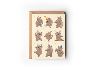 Dancing Wombats Card by 9to5 Studios, cream A6 birthday greeting card with nine brown wombats in colorful party hats and gold stars, shown with kraft envelope